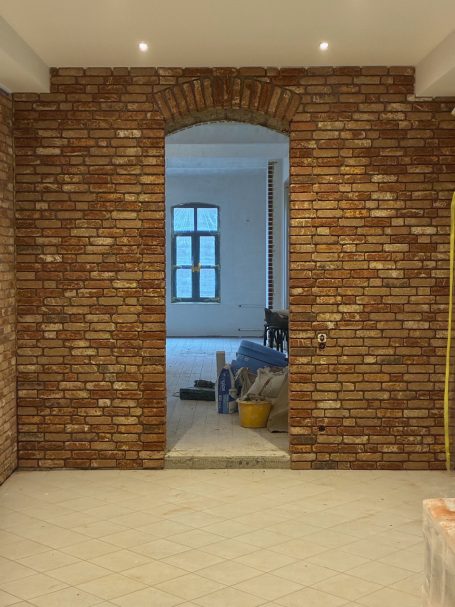 Wand mit Klinkerriemchen mit einem Torbogen, Durchgang zu einem hellen Raum im Hintergrund.